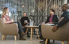 2021/10/conversation-245.jpg Happy young people sitting together in office discussing business. Office workers having a meeting in lobby. Diverse business people at startup.