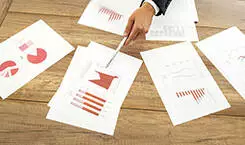 2021/10/data-choosing-245.jpg Businessman giving a presentation with assorted analytical graphs and charts spread out on the desk in front of him pointing to relevant information with a pen.