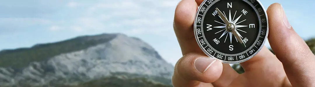  2021/10/landscape-compass-header.jpg Close up of the hand of a man holding a magnetic compass over a landscape view as he uses it to navigate when exploring or travelling in the countryside