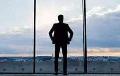 2021/10/mature-businessman-standing-at-the-window-245.jpg Mature businessman standing at the big windows in a hotel. Man looking at city in the evening. Rear view.