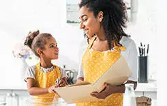  2021/10/mom-and-daughter-245.jpg african american mother holding cookbook and daughter preparing dough in kitchen, looking at each other