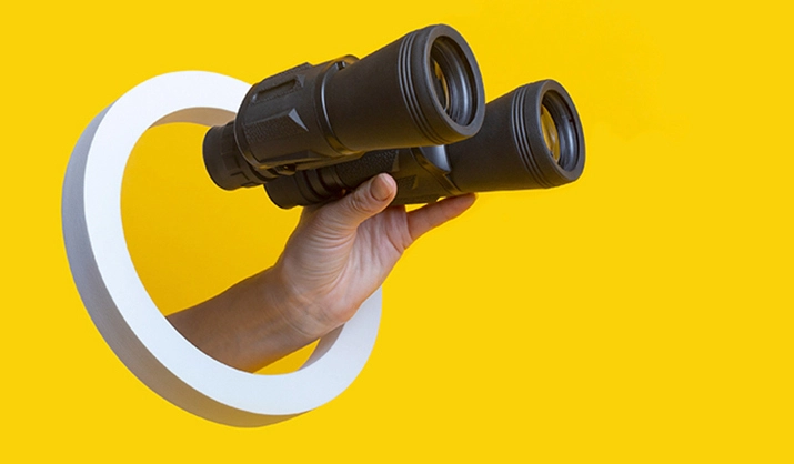 Female hand holding binoculars through a hole on a bright yellow background
