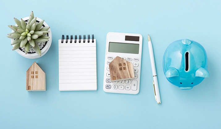 Calculator with house model, notebook and piggy bank, housing finance concept on blue table, top view